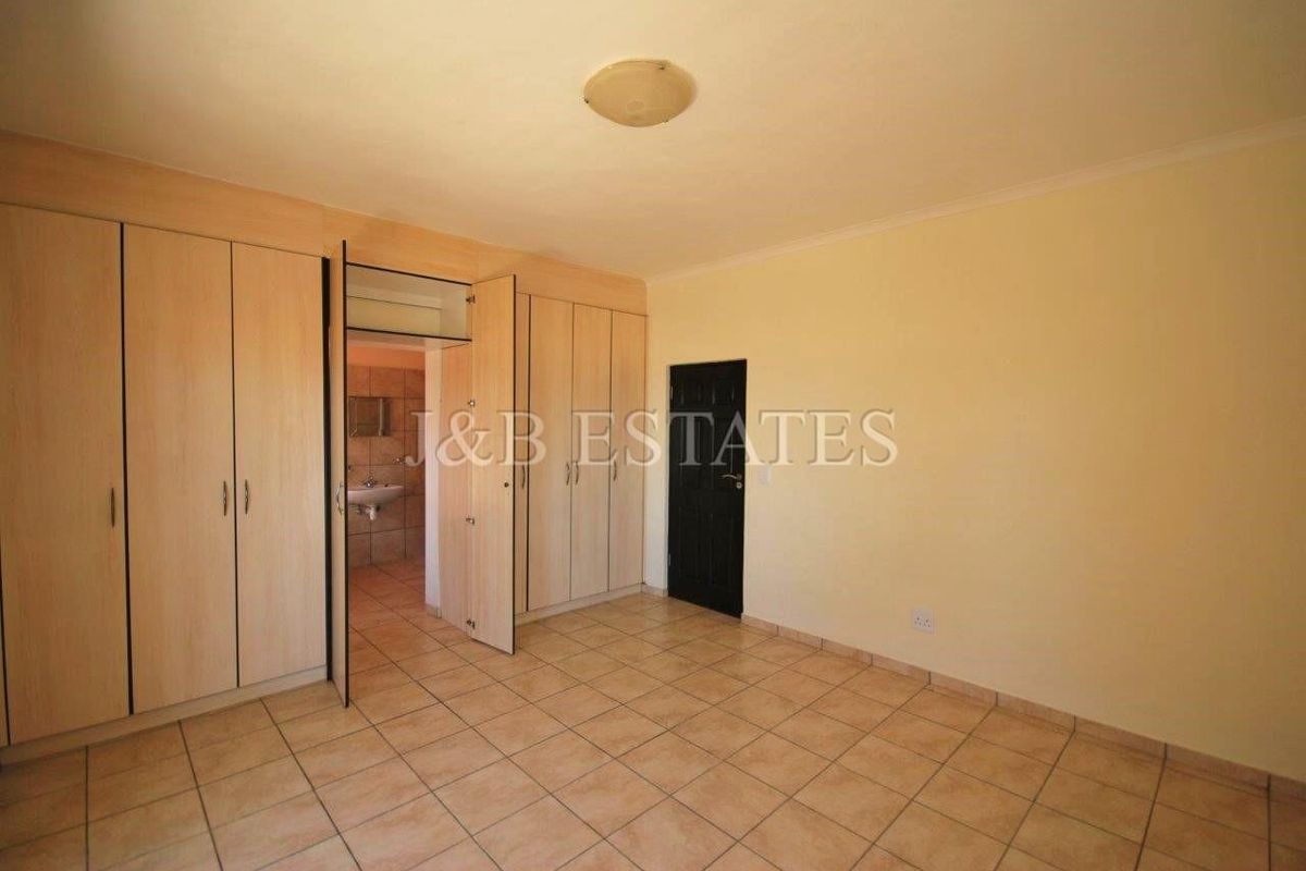 A spacious, empty bedroom featuring neutral tiled flooring and extensive built-in wooden cupboards, with an adjoining view into a tiled bathroom area.