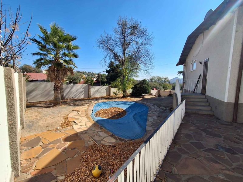 Rear outdoor area featuring a swimming pool with cover, stone paved patio, and boundary wall, requiring maintenance and landscaping improvements.