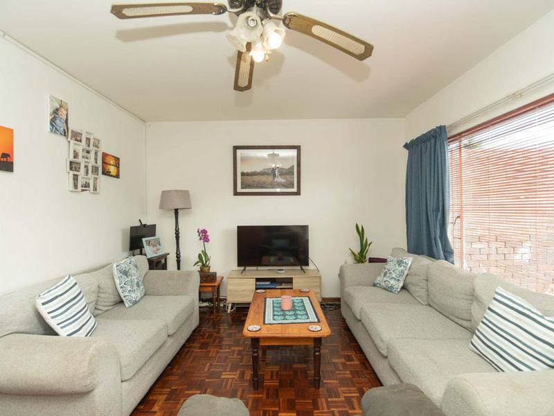 A comfortable living room featuring classic parquet flooring, a ceiling fan, and large windows with venetian blinds, offering a warm and inviting atmosphere.