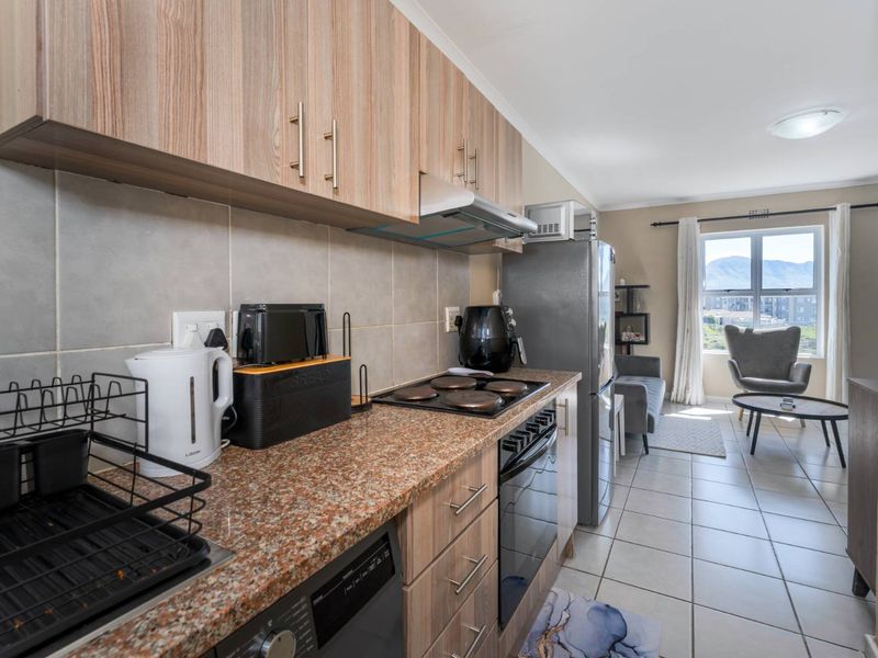 An open-plan kitchen and living area featuring wood-finish cabinetry, granite countertops, and tiled flooring, leading to a bright living space with mountain views.