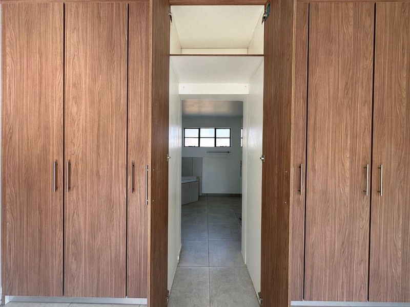 A clean, empty bedroom featuring modern wood-finish built-in cupboards and tiled flooring, with a direct view into an adjoining bathroom area.