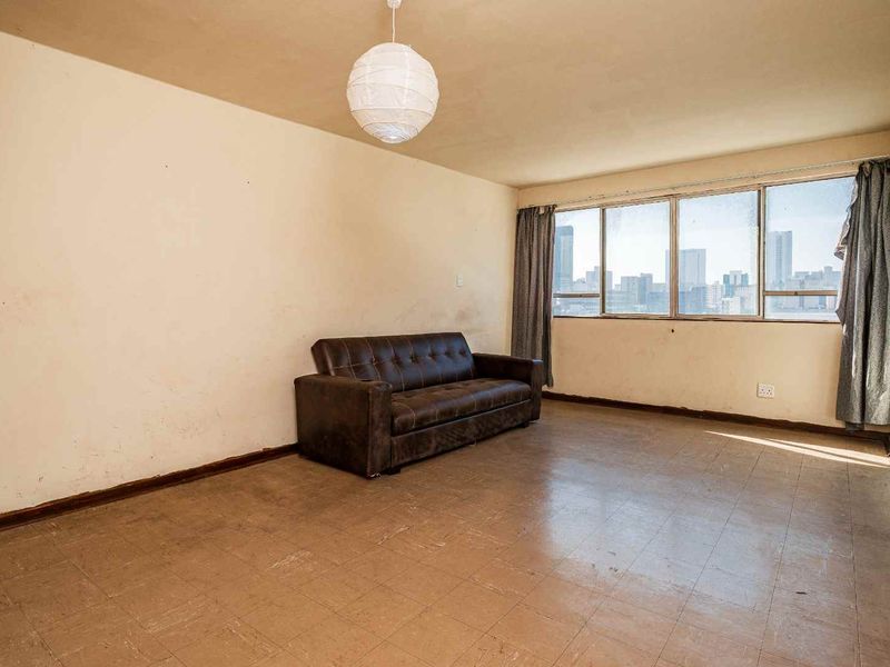 A spacious living room featuring large windows with city views, tiled flooring, and neutral walls in need of cosmetic refreshment.