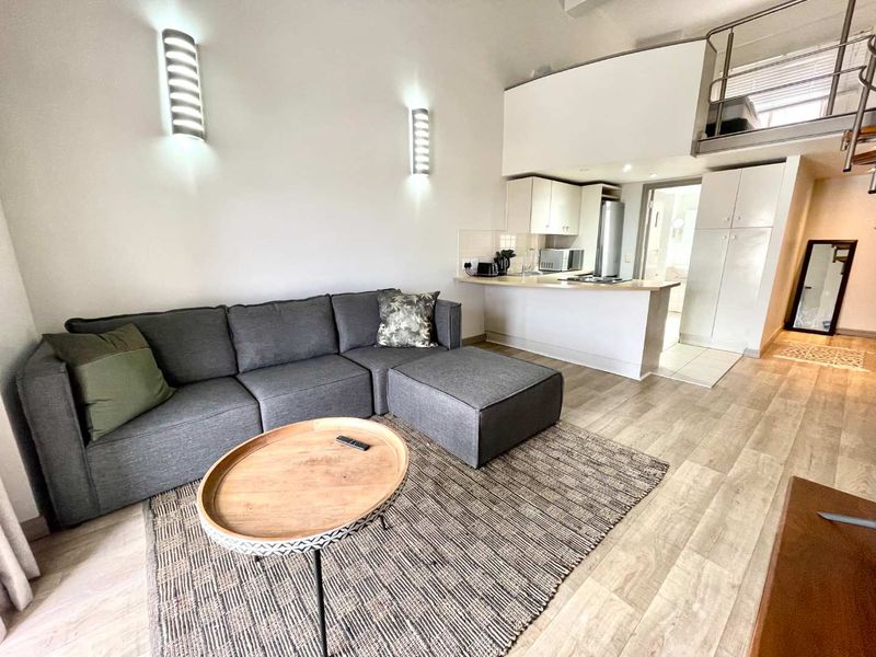 Open-plan living area featuring laminate flooring, a compact kitchen with built-in cabinetry, and a mezzanine level with metal staircase railing.