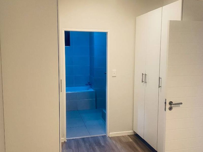 A clean, modern hallway featuring light-toned laminate flooring and ample built-in cupboard storage, leading directly into a tiled bathroom area.