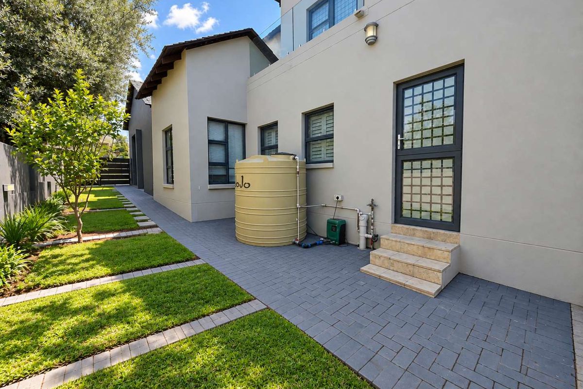 Rear exterior view featuring a paved walkway, landscaped garden, and a water storage tank installed against the building facade.
