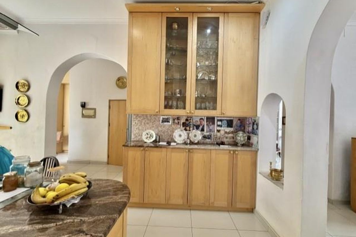 A kitchen area featuring light wood built-in cabinetry with glass display sections, stone countertops, and a tiled backsplash, framed by architectural archways.