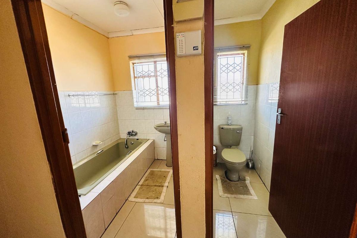 A dated bathroom featuring a built-in bathtub, pedestal basin, and separate toilet area with tiled finishes and security bars on the windows.