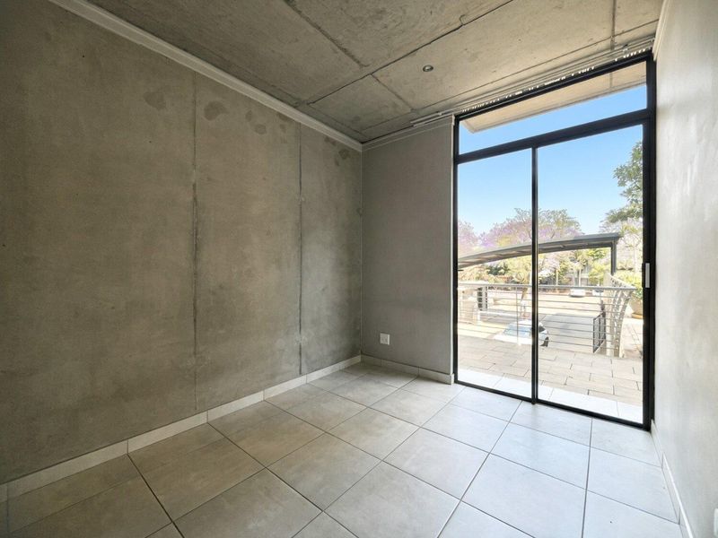 An empty bedroom featuring an industrial aesthetic with exposed concrete walls and ceiling, tiled flooring, and a large sliding glass door leading to a balcony.