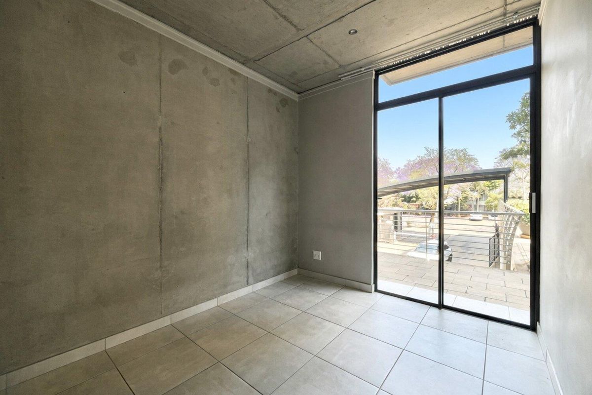 An empty bedroom featuring an industrial aesthetic with exposed concrete walls and ceiling, tiled flooring, and a large sliding glass door leading to a balcony.