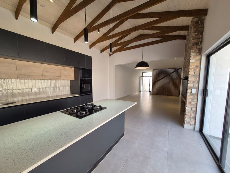 Modern open-plan kitchen and living area featuring exposed timber beams, a central island with gas hob, and an exposed brick fireplace.