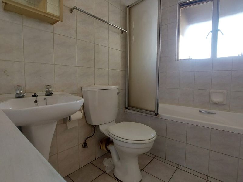 A dated bathroom featuring a pedestal basin, toilet, and a combined bathtub with a shower screen, finished with neutral wall and floor tiles.