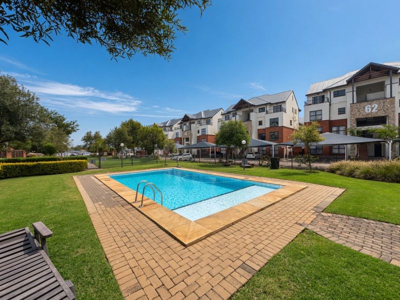 A well-maintained communal swimming pool area featuring paved surrounds, manicured lawns, and landscaped gardens within a secure apartment complex.