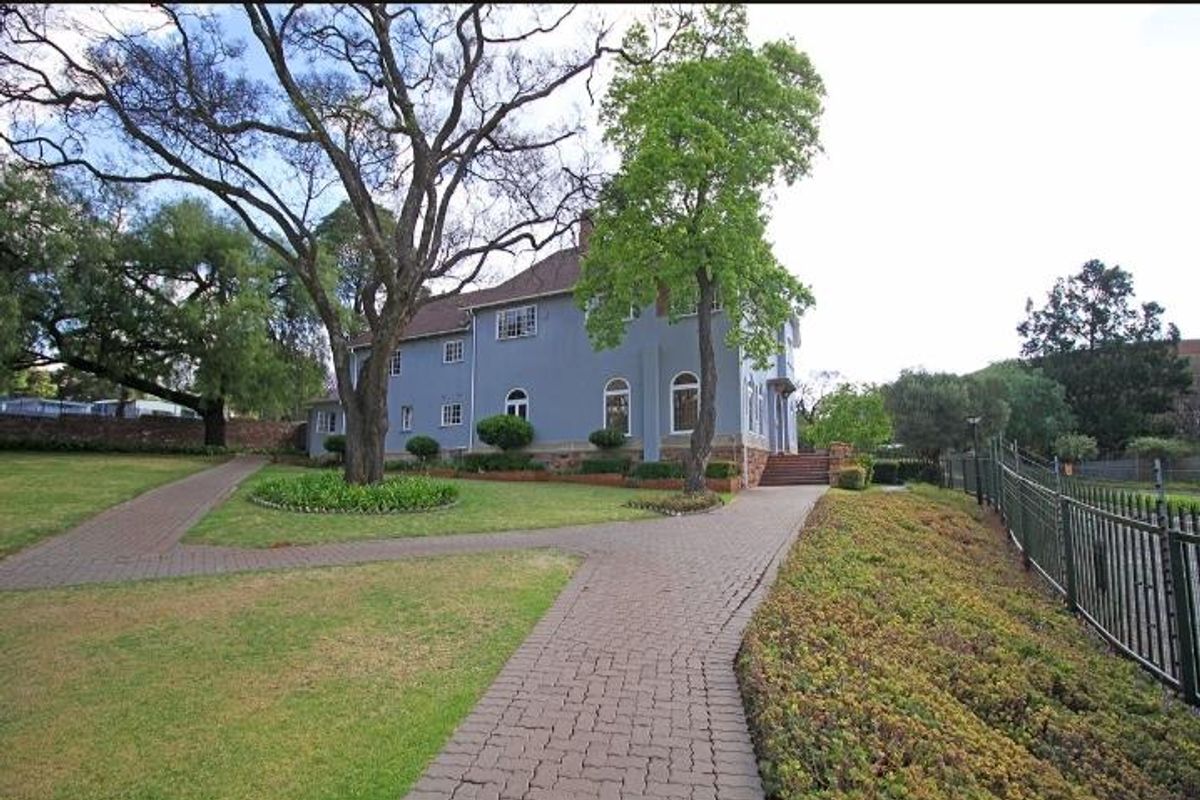 Exterior view of the apartment building featuring a paved walkway, landscaped garden, mature trees, and a secure perimeter fence.