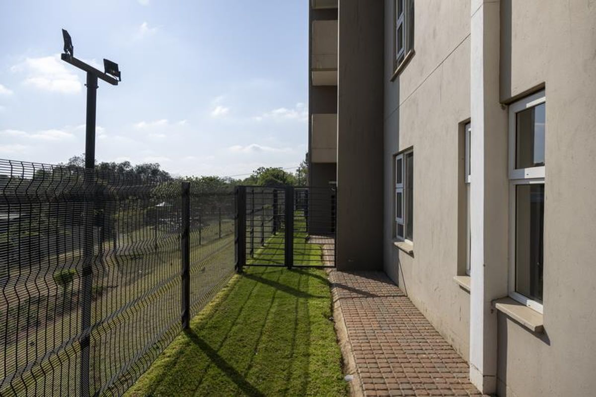 Exterior view of the apartment building showing a paved walkway, manicured lawn, and secure perimeter fencing.