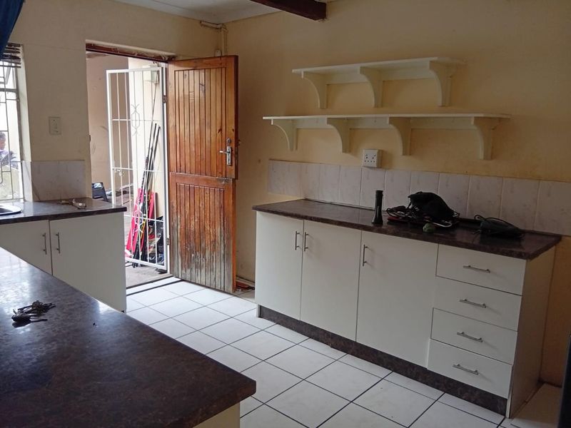 A functional kitchen area featuring white cabinetry, dark laminate countertops, tiled flooring, and wall-mounted shelving, with direct access to an exterior space.