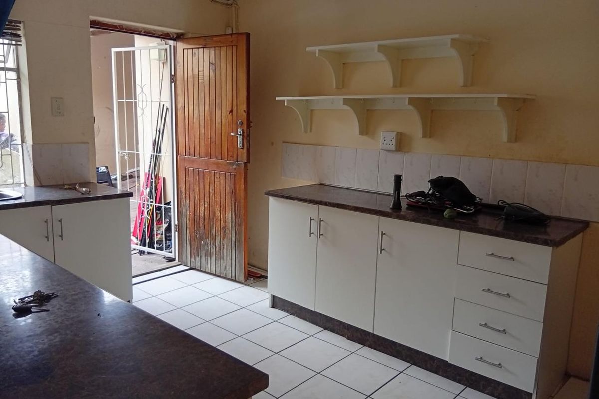 A functional kitchen area featuring white cabinetry, dark laminate countertops, tiled flooring, and wall-mounted shelving, with direct access to an exterior space.