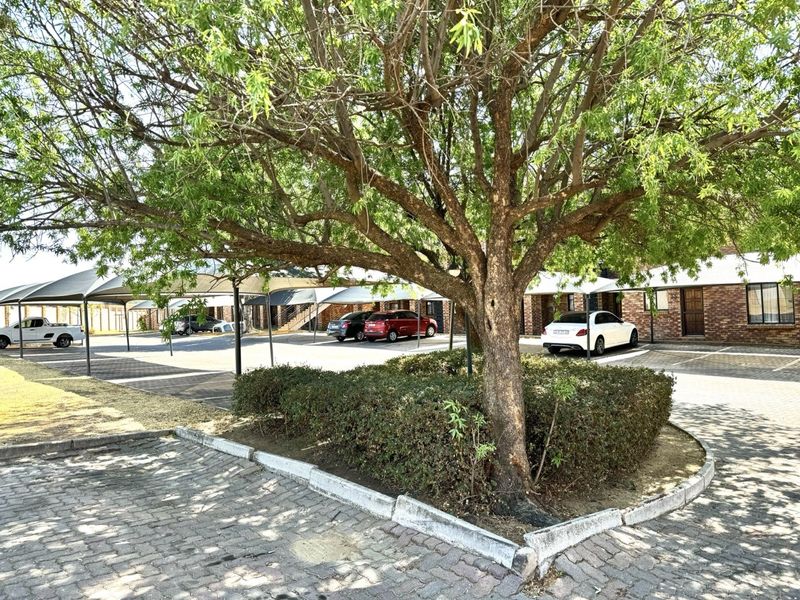 Exterior view of the apartment complex featuring a paved parking area with covered carports, brick building facades, and established garden landscaping.