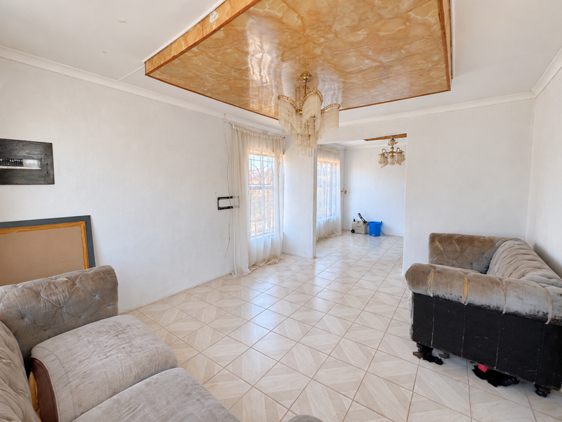 Spacious living area featuring tiled flooring, a decorative ceiling panel with a crystal chandelier, and large windows with security bars.