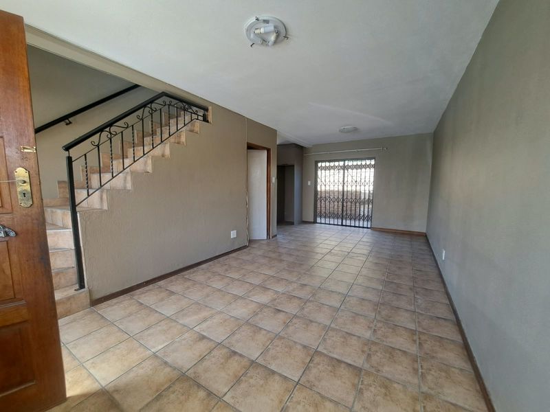 An empty, open-plan living area featuring tiled flooring, a staircase with a wrought iron balustrade, and a security gate leading to the exterior.