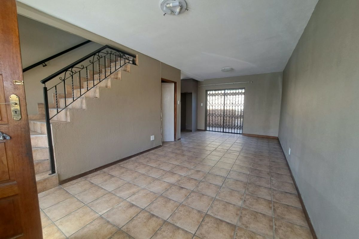An empty, open-plan living area featuring tiled flooring, a staircase with a wrought iron balustrade, and a security gate leading to the exterior.