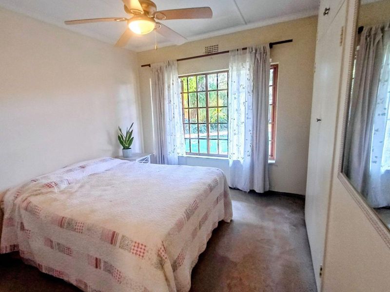 A bedroom featuring built-in cupboards, a ceiling fan, and a window with security bars overlooking the garden.