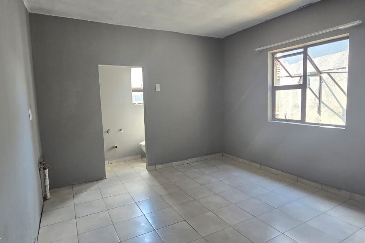 An empty bedroom featuring tiled flooring, neutral grey walls, and an adjoining small utility or bathroom space with visible plumbing connections.