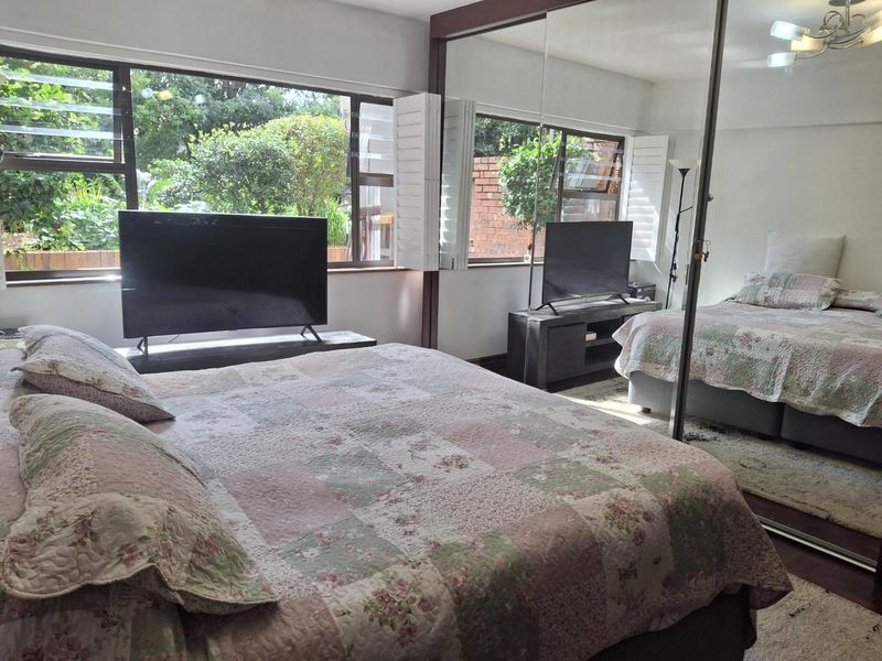 A bedroom featuring large mirrored sliding wardrobe doors and expansive windows with louvre shutters, providing natural light and garden views.