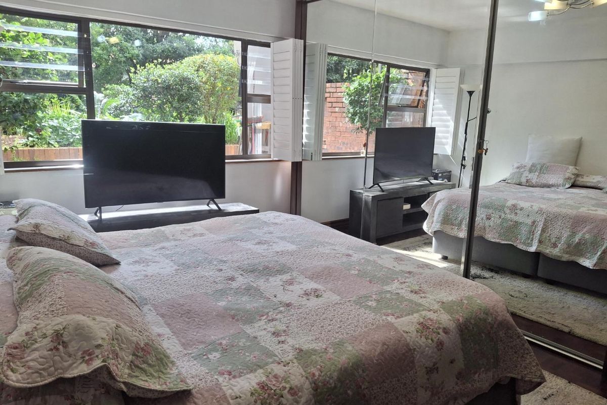 A bedroom featuring large mirrored sliding wardrobe doors and expansive windows with louvre shutters, providing natural light and garden views.