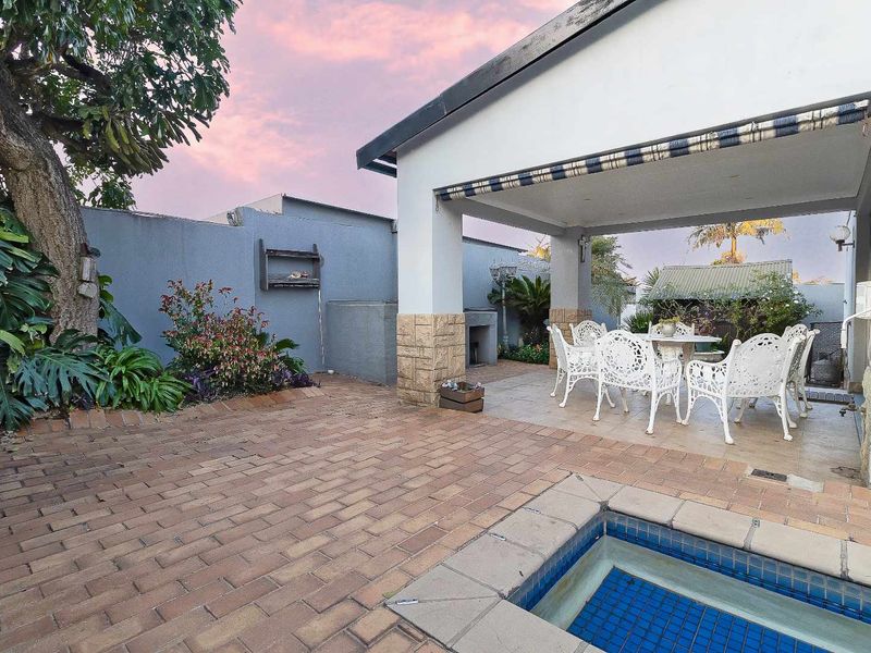 A covered patio area featuring a built-in braai, paved flooring, and direct access to a swimming pool, set within a private walled garden.