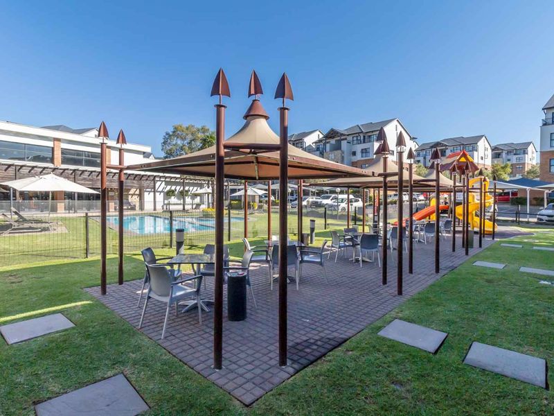Communal outdoor area featuring a swimming pool, paved patio with shade structures, landscaped lawn, and playground equipment within a residential apartment complex.