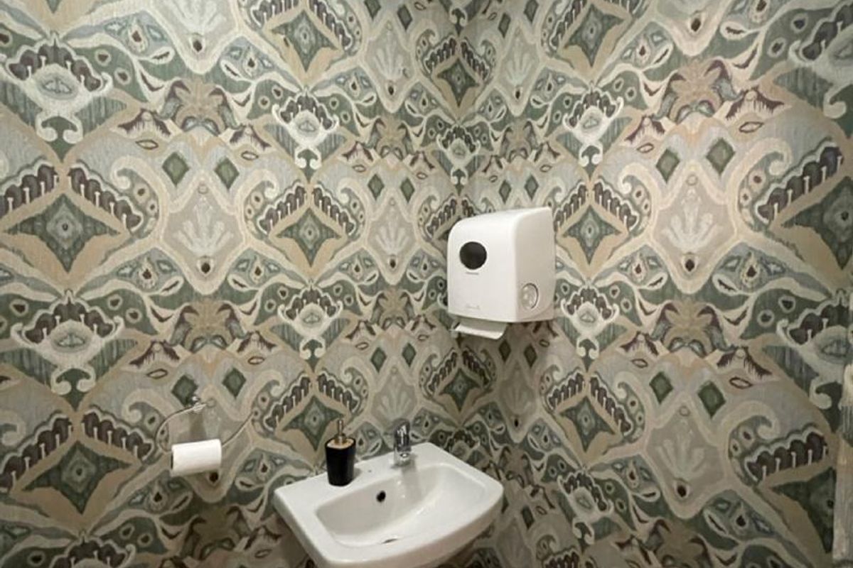 A compact restroom featuring patterned wallpaper, a wall-mounted ceramic basin, and a wall-mounted paper towel dispenser.