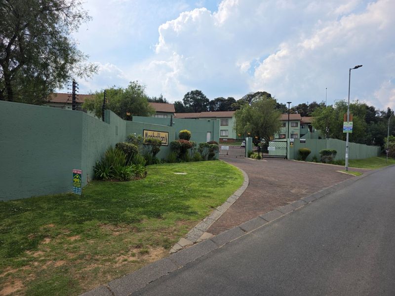 Street view of the complex entrance featuring a paved driveway, secure gated access, perimeter wall with electric fencing, and well-maintained garden area.