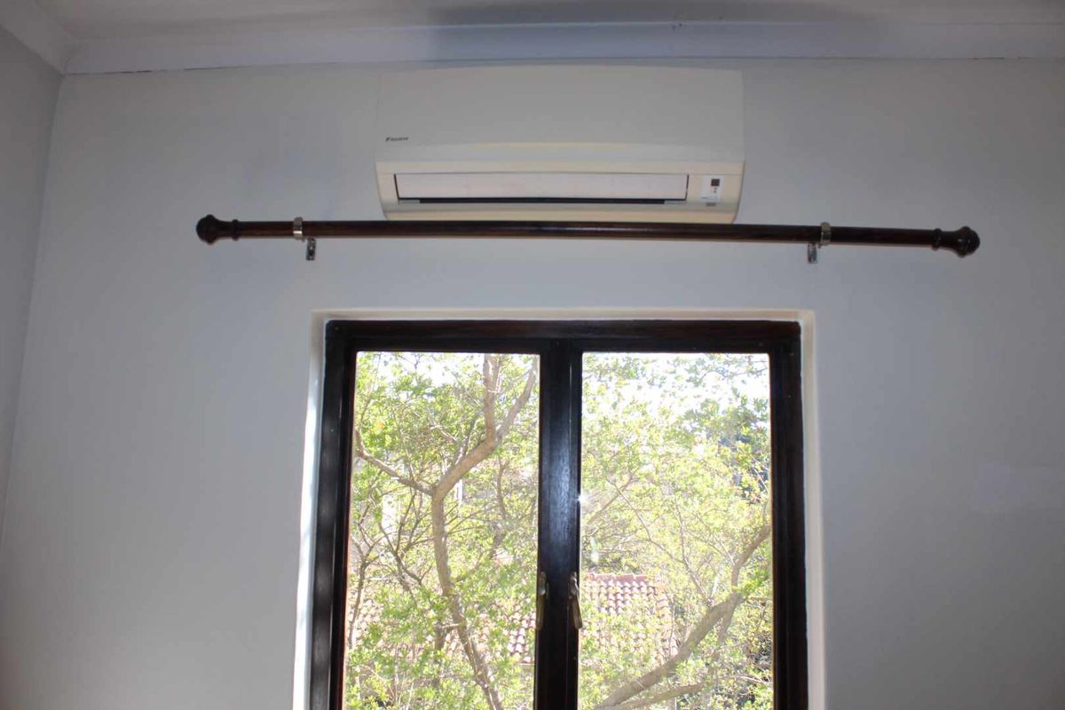 A bedroom interior featuring a wall-mounted air conditioning unit, a timber-framed window, and a curtain rod.