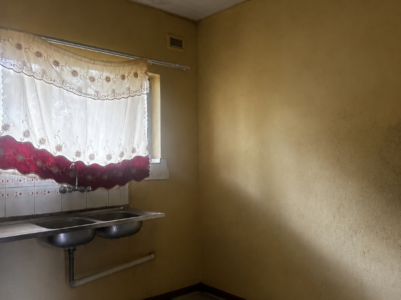A basic, empty kitchen area featuring a double stainless steel sink, tiled splashback, and checkerboard flooring, requiring cosmetic updates and modernization.
