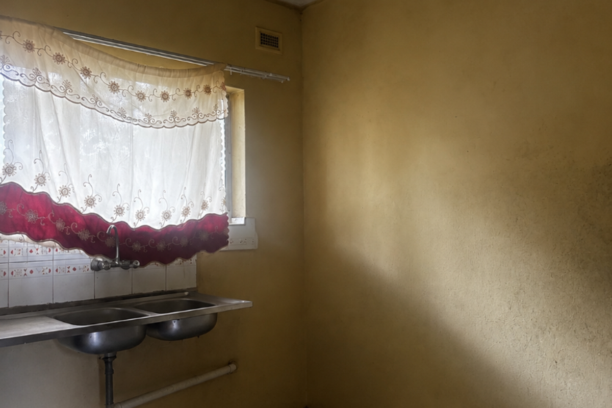 A basic, empty kitchen area featuring a double stainless steel sink, tiled splashback, and checkerboard flooring, requiring cosmetic updates and modernization.
