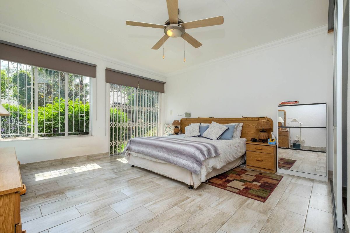 Spacious bedroom featuring light-toned tiled flooring, a ceiling fan, and large windows with security bars, providing direct access to the exterior.