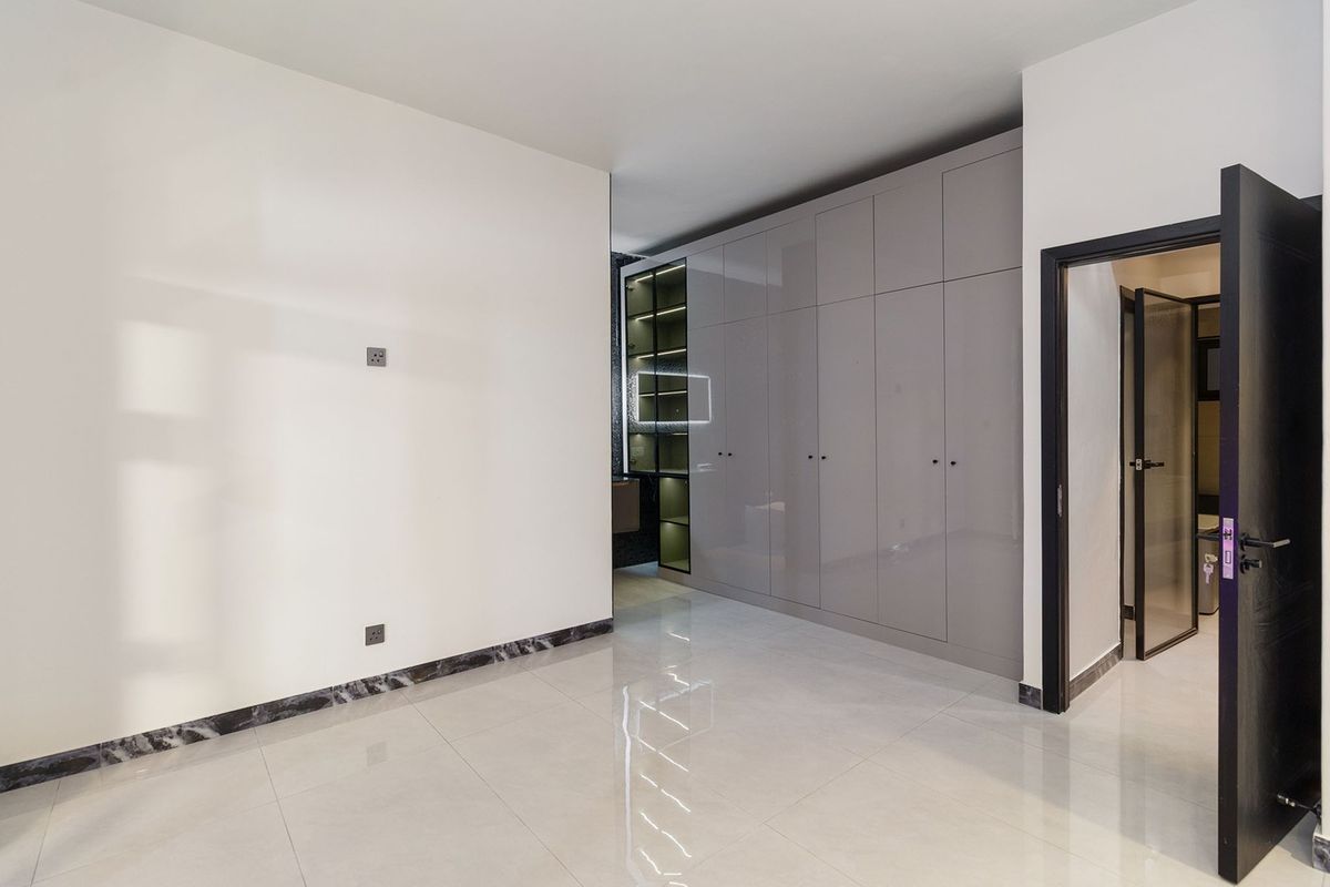 A modern, empty bedroom featuring high-gloss built-in wardrobes, sleek tiled flooring, marble skirting, and an integrated display shelving unit.
