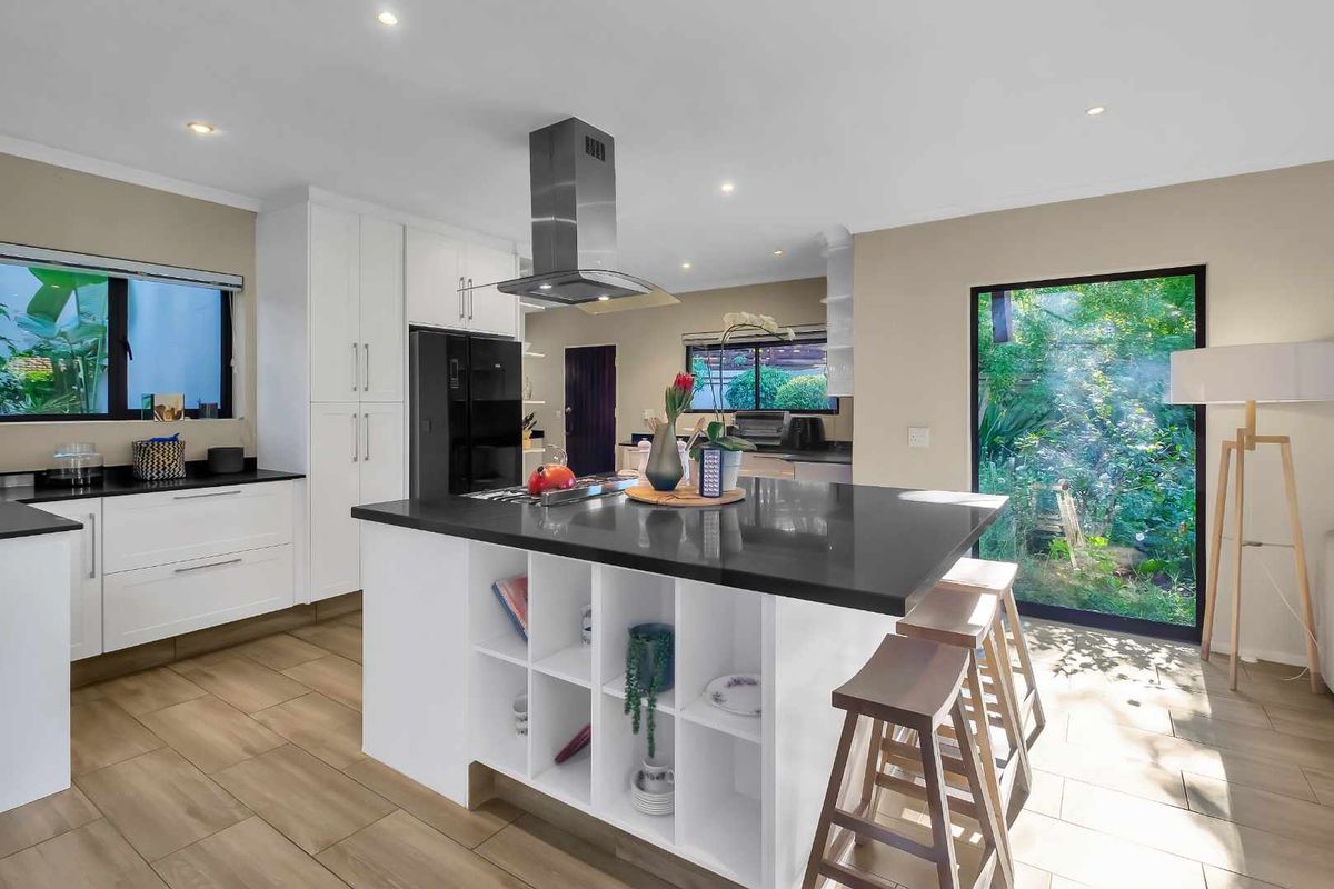 Modern kitchen featuring a central island with stone countertops, white cabinetry, stainless steel extractor fan, and large windows overlooking the garden.