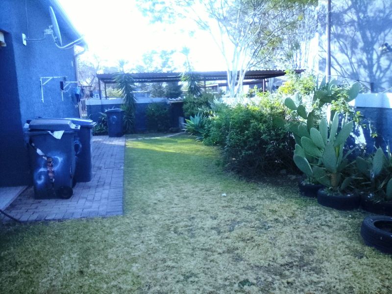 A rear garden area featuring a paved walkway, lawn, and boundary wall with an adjacent carport structure.