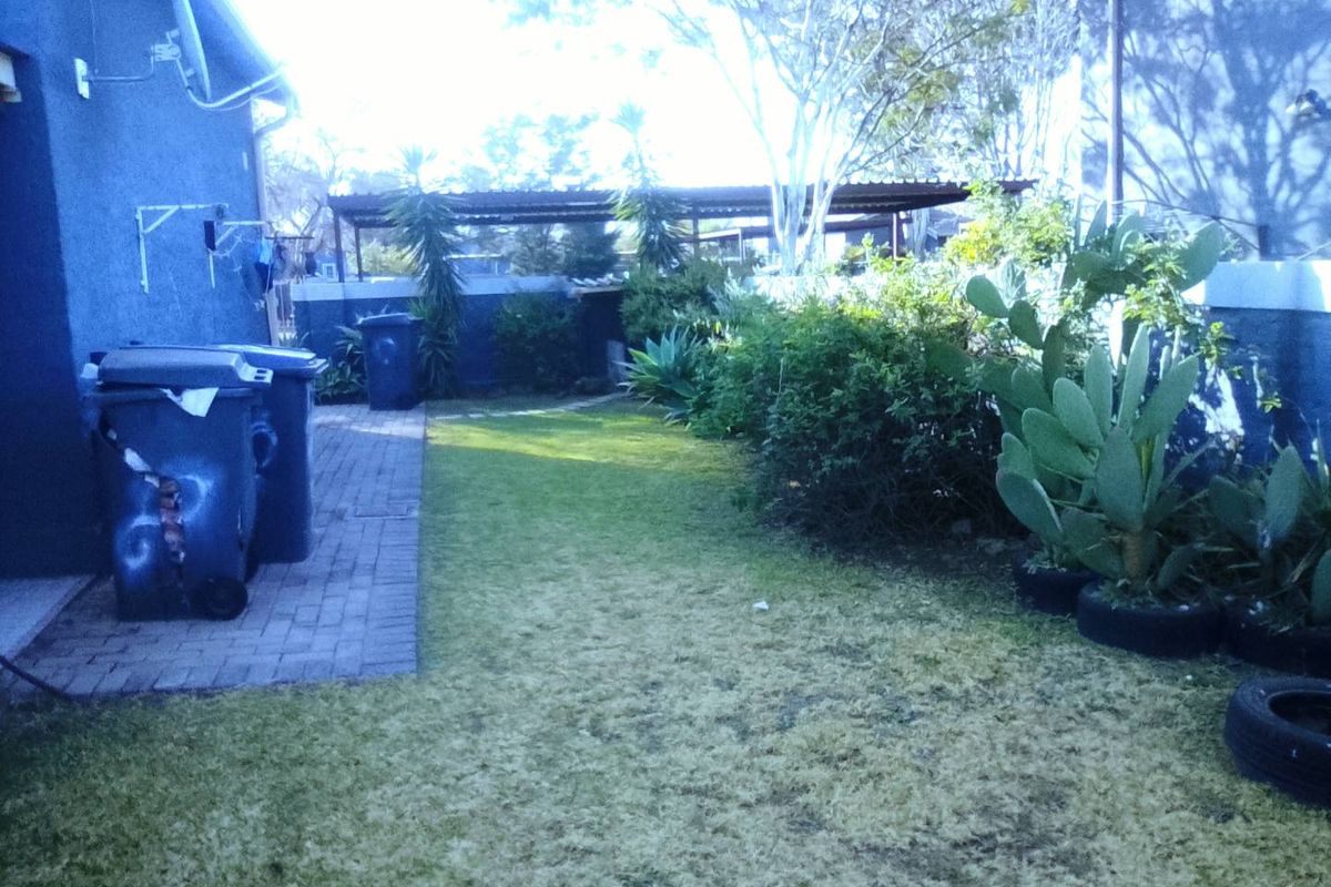 A rear garden area featuring a paved walkway, lawn, and boundary wall with an adjacent carport structure.