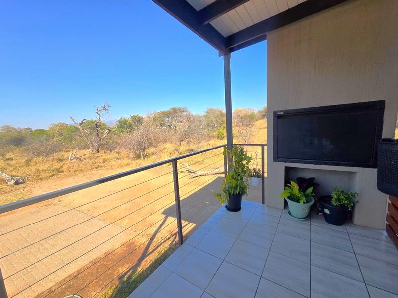 A covered outdoor patio featuring tiled flooring, a built-in braai, and metal railings, offering expansive views of the surrounding natural landscape.