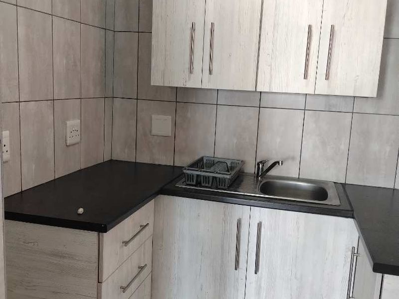 A functional scullery area featuring light wood-grain cabinetry, dark laminate countertops, a stainless steel sink, and neutral wall and floor tiling.