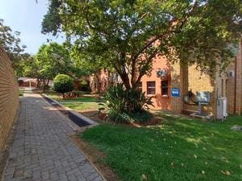 A view of the apartment complex exterior featuring a brick facade, a paved walkway, and well-maintained garden areas with mature trees.