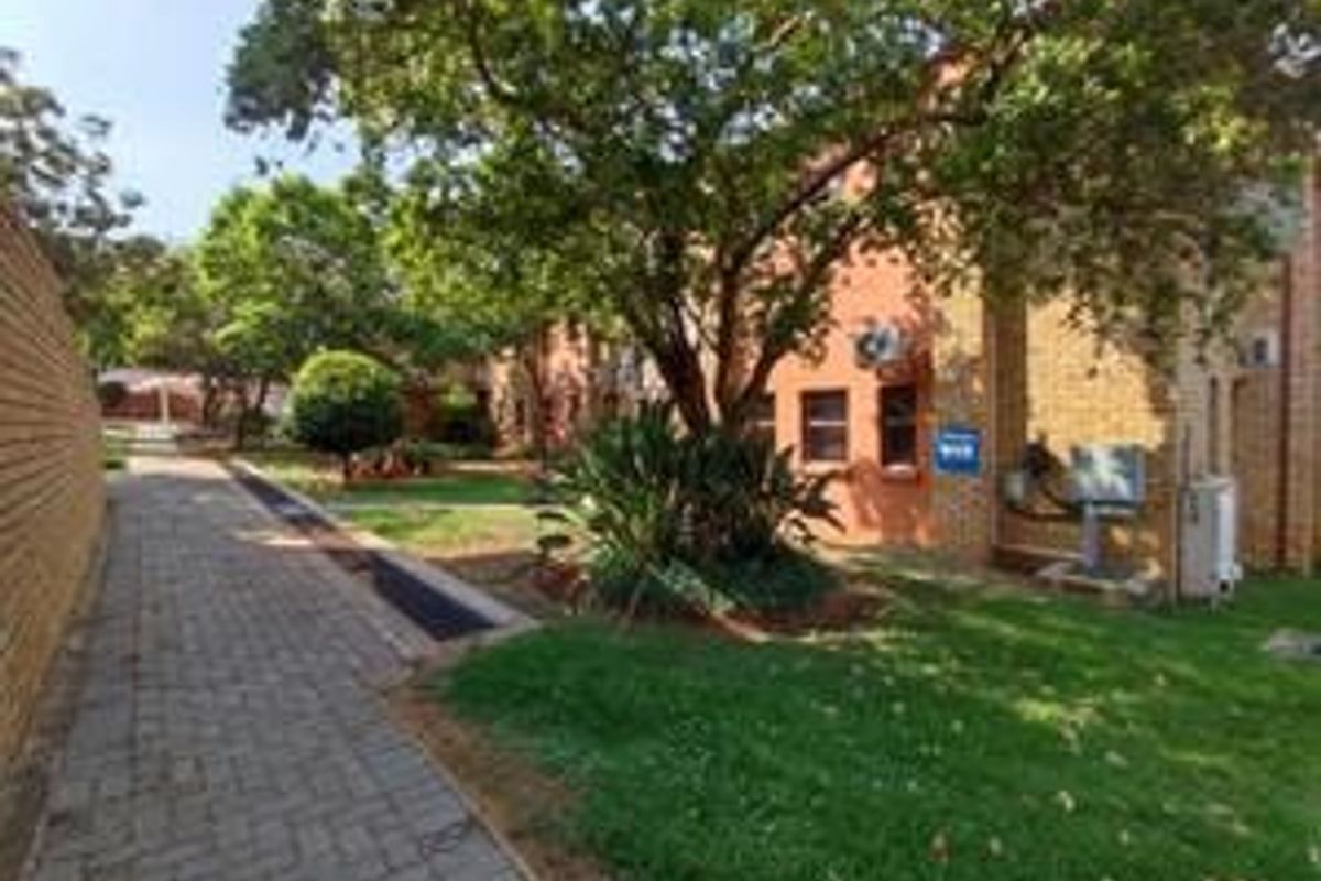 A view of the apartment complex exterior featuring a brick facade, a paved walkway, and well-maintained garden areas with mature trees.