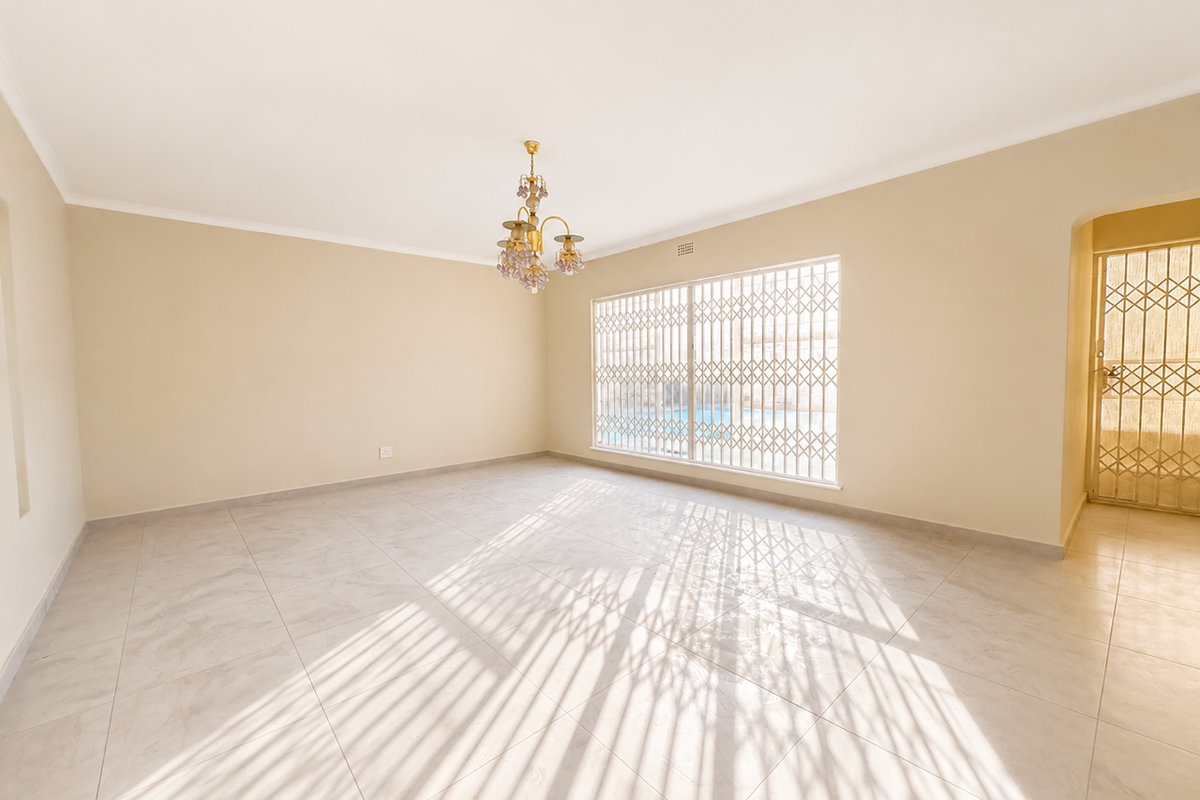 A bright, empty living room featuring neutral tiled flooring, a decorative chandelier, and secure window and door grilles.