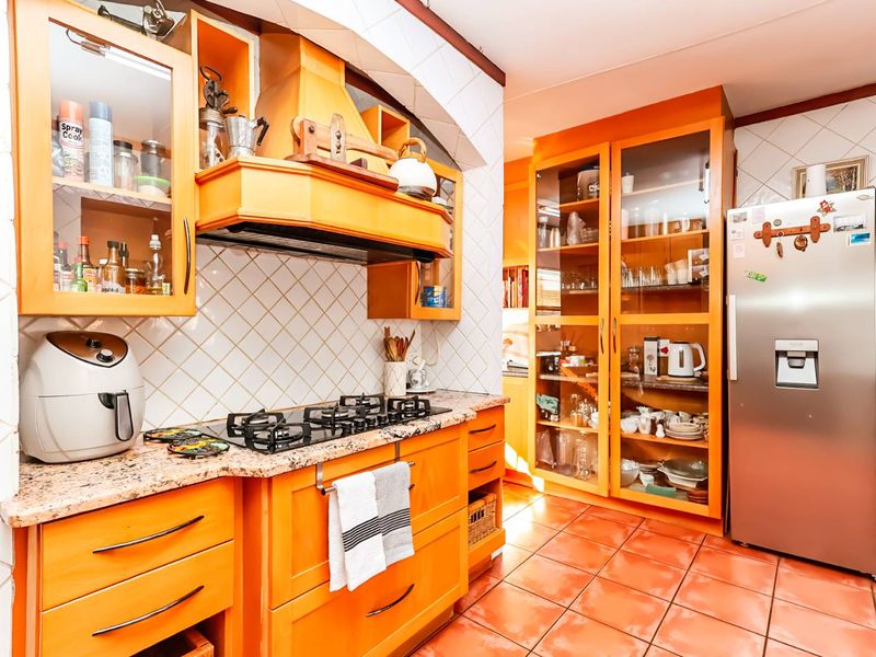 A functional kitchen featuring vibrant orange cabinetry, granite countertops, a gas hob with an extractor fan, and ample glass-fronted storage space.