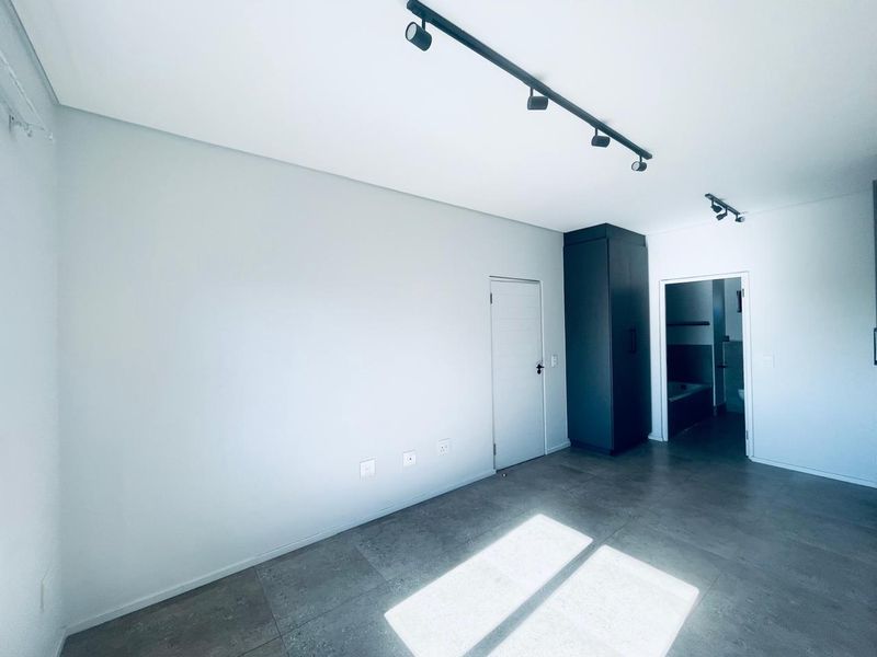A modern, empty bedroom featuring sleek tiled flooring, contemporary track lighting, and a built-in cupboard, with an adjoining bathroom visible through the doorway.
