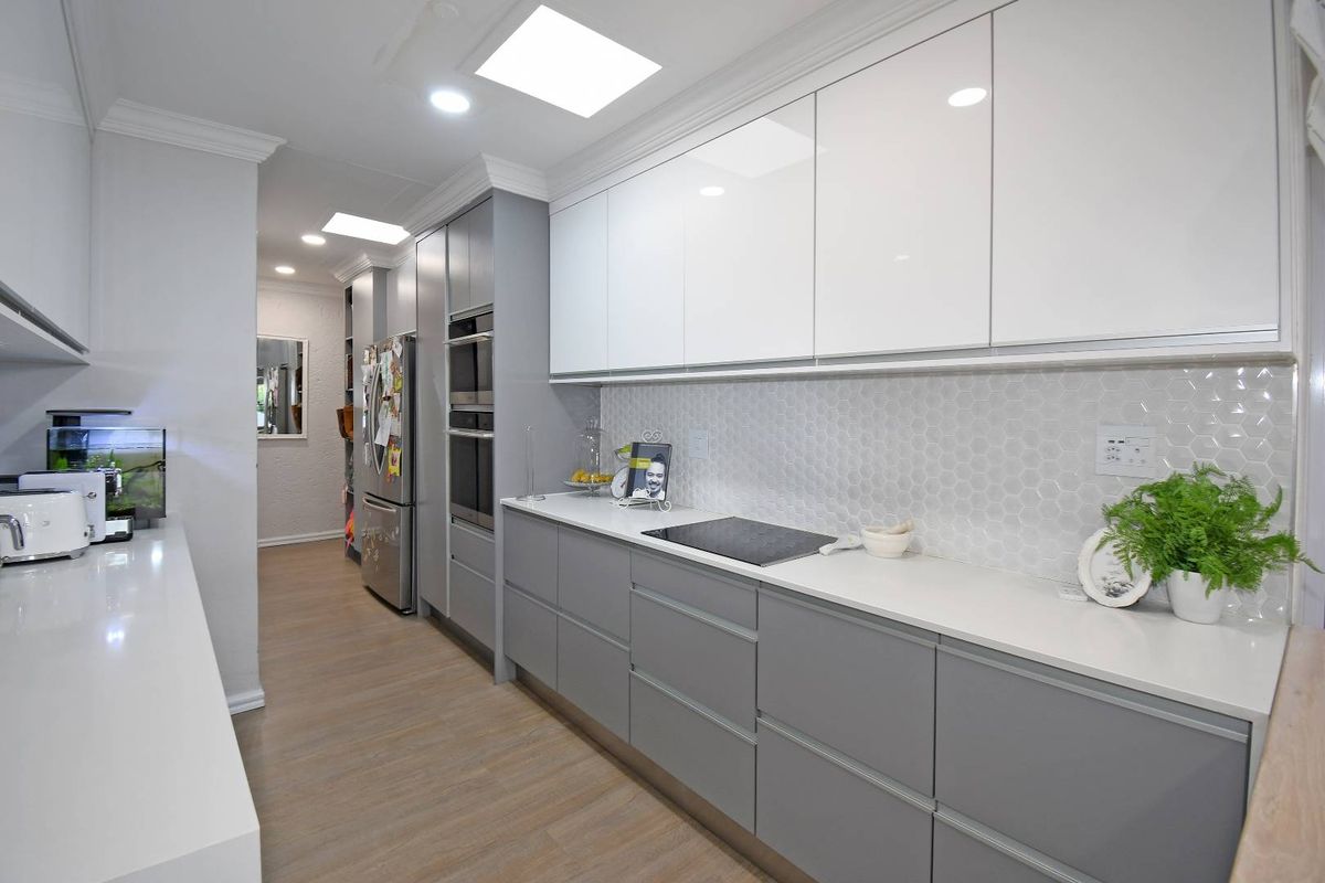 Modern galley-style kitchen featuring sleek grey and white cabinetry, stone countertops, integrated ovens, a glass cooktop, and a stylish hexagonal tile backsplash.