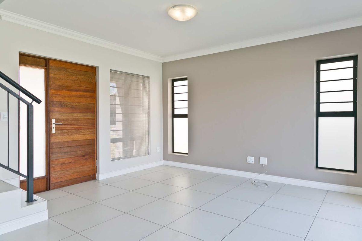A bright, empty living area featuring neutral tiled flooring, a solid wooden front door, and modern aluminium window frames.