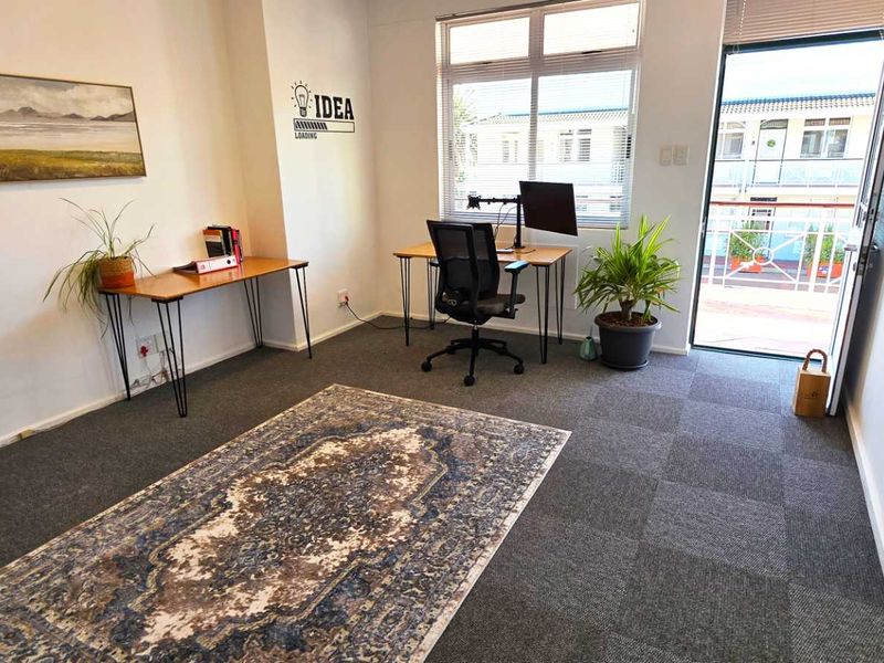 A bright office space featuring carpet tile flooring, large windows for natural light, and direct access to an outdoor balcony area.